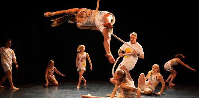 Sirkusryhmä lavalla, yksi roikkuu narun varassa, muut istuvat tai seisovat lattialla