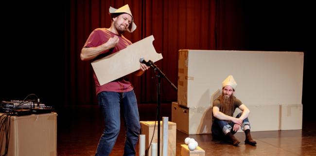 Mies soittaa puupalasta hattu päässä, toinen katselee taustalla