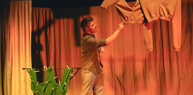 Lavalla seisova henkilö osoittaa sormellaan ylhäältä roikkuvaa vaatekappaletta. Taustalla on lämpimän oranssiksi valaistut verhot. Sivulla näkyy pyykinkuivausteline, jossa vihreitä vaatteita on kiinnitetty pyykkipojilla. Tunnelma on teatterimainen ja harkitusti lavastettu.