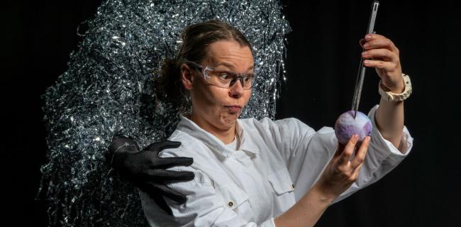 Suojalaseja ja valkoista työasua käyttävä henkilö tutkii käsissään olevaa violettia pallomaista esinettä metallisella työkalulla; taustalla näkyy tumma hahmo ja hopeanhohtoinen, sirpaleinen pinta.