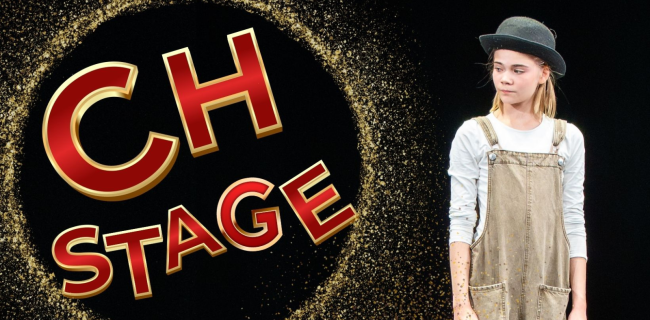 Mustalla taustalla kultaisista hiukkasista muodostuva rengas ja sen sisällä punakultainen teksti ‘CH STAGE’. Oikealla puolella seisoo nuori henkilö, jolla on harmaa hattu, vaalea paita ja haalarit, ja hän katsoo sivulle.