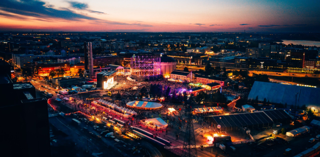 Aerial view of Flow Festival at dusk in Helsinki. The festival grounds, set within a former industrial area, glow with colorful lights as large crowds gather around the main stage and numerous tents, food stalls, and event spaces. In the background, the illuminated city skyline stretches beneath a sunset sky painted in soft pink and blue hues.