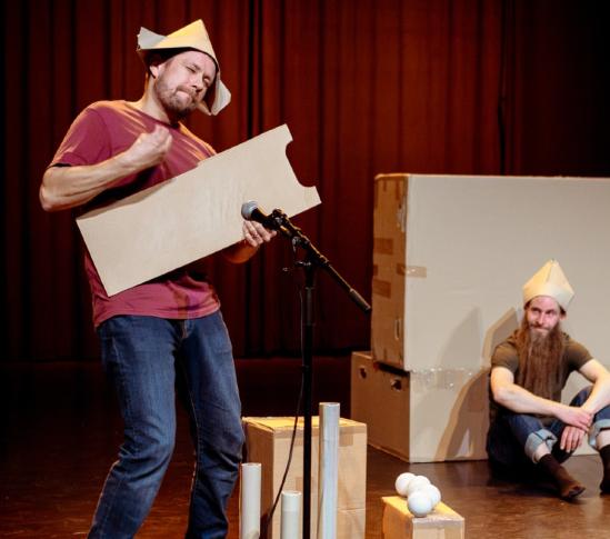 Mies soittaa puupalasta hattu päässä, toinen katselee taustalla