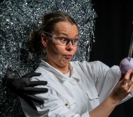A person wearing safety goggles and a white work outfit examines a small purple spherical object with a metal tool; behind them is a dark figure and a shimmering, fragmented silver background.