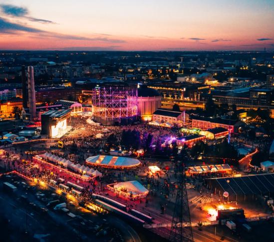 Ilmakuva Flow Festivalista iltahämärässä Helsingissä. Entisen teollisuusalueen keskelle rakentunut festivaalialue hehkuu värikkäissä valoissa, ja suuri yleisö on kerääntynyt päälavan sekä lukuisten telttojen ja tapahtumapisteiden ympärille. Taustalla näkyy kaupungin valaistu siluetti ja auringonlaskun sävyttämä taivas.